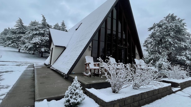 Une maison moderne avec un toit en pente, de grandes fenêtres et un jardin enneigé agrémenté d’un petit arbre et d’un banc.