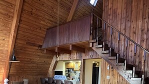 L’intérieur d’un chalet rustique, avec un escalier en bois, deux canapés ornés de coussins décoratifs, un coin repas et une cuisine visible à travers une porte ouverte.