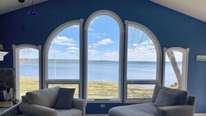 Ein Zimmer mit einem großen Bogenfenster mit Blick auf ein Gewässer, zwei Sesseln und einem Kamin aus Stein.