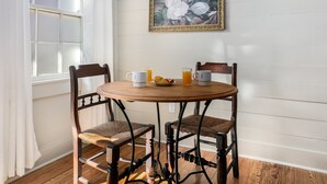 Een eetgedeelte met een ronde houten tafel, twee gestoffeerde stoelen, een bloemschilderij in een lijst aan de muur, en een raam met gordijnen.