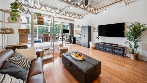 Ampla sala de estar com sofá, apoio para os pés e mesa de jantar com frutas. Há uma TV na parede, um ventilador de teto e uma janela grande que proporciona iluminação natural.