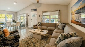 A spacious living room with a large sofa, a coffee table, and a ceiling fan. There is a dining area with a table and chairs, and a painting on the wall. - Historic Downtown - close to beaches - firepit - playground - pet friendly (St. Augustine)