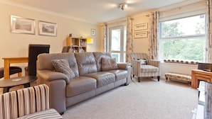 A living room with a grey sofa, striped armchair, and a small wooden table. There is a bookshelf with books and decorative items, and a window with curtains. - Herdwick Place, spacious cottage for 4 in spectacular Langdale Valley (Great Langdale)