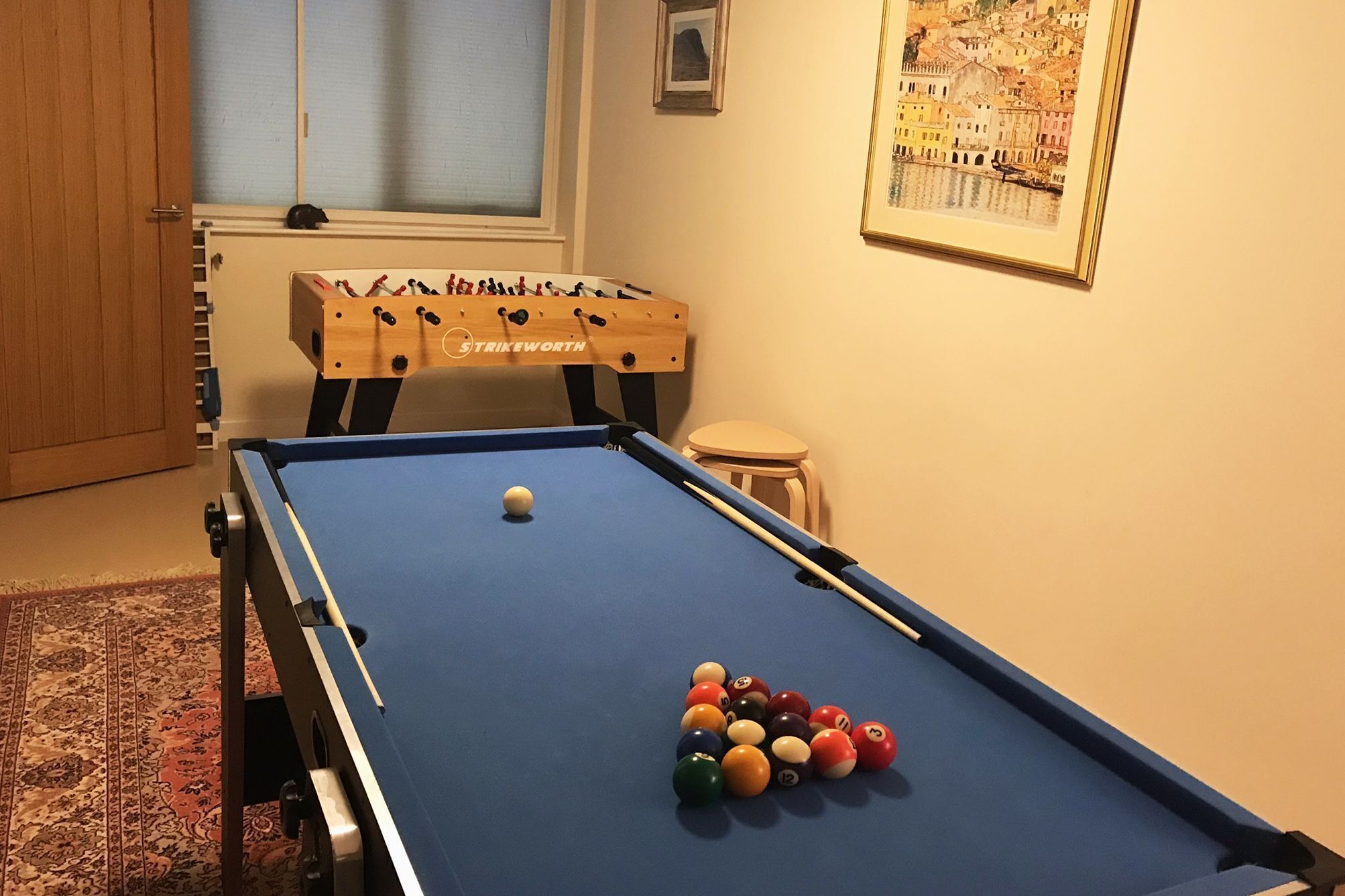Ein Billardtisch mit Billardkugeln, ein Tischfußballspiel und ein gerahmtes Stadtbild.