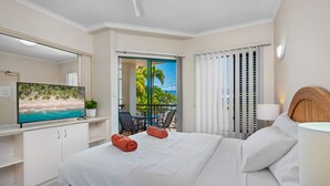 A hotel room with a bed, a TV, a balcony with a view of palm trees and the ocean, and a desk with a plant. - Tropic Towers: Cairns City Family Apartment (Cairns North)
