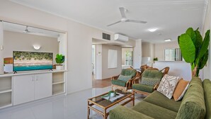 A spacious living room with a flat-screen TV, a coffee table, and a sofa. There are ceiling fans, a kitchen area, and a large plant. - Tropic Towers: Cairns City Family Apartment (Cairns North)