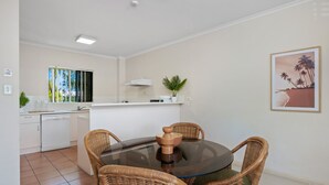 A compact kitchen with white cabinets, a dining table with four wicker chairs, and a potted plant on the counter. - Tropic Towers: Cairns City Family Apartment (Cairns North)