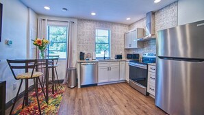 A compact kitchen with a dishwasher, microwave, stove, and refrigerator. There is a dining area with a table and chairs, and a window with blinds. - 1930s Cape Cod Bungalow at Firmstone Manor (Clifton Forge)