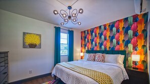A bedroom with a bed, a chandelier, a colorful wall mural, a window with blue curtains, and a dresser. - 1930s Cape Cod Bungalow at Firmstone Manor (Clifton Forge)