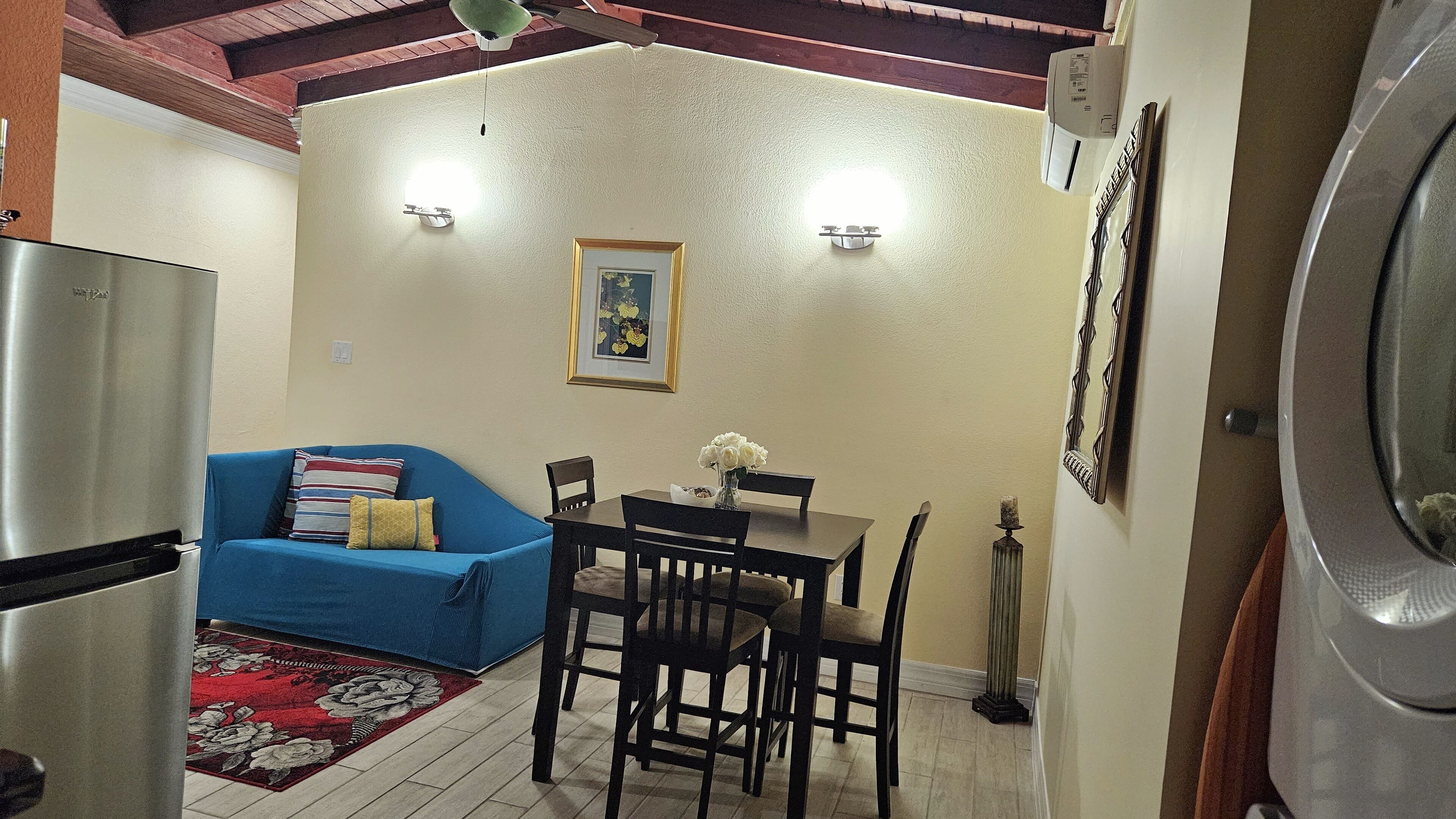 Un espace de vie compact comprenant un coin cuisine, un coin repas avec une table et des chaises, un canapé bleu avec des coussins et un tapis rouge à motifs floraux.