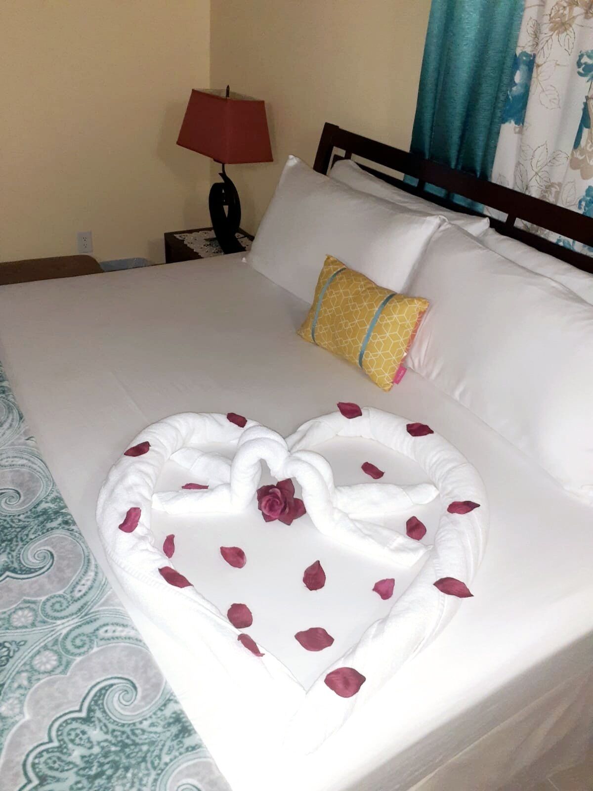 Cama bem arrumada com lençóis brancos, arranjo de toalhas em formato de coração e uma única almofada decorativa.