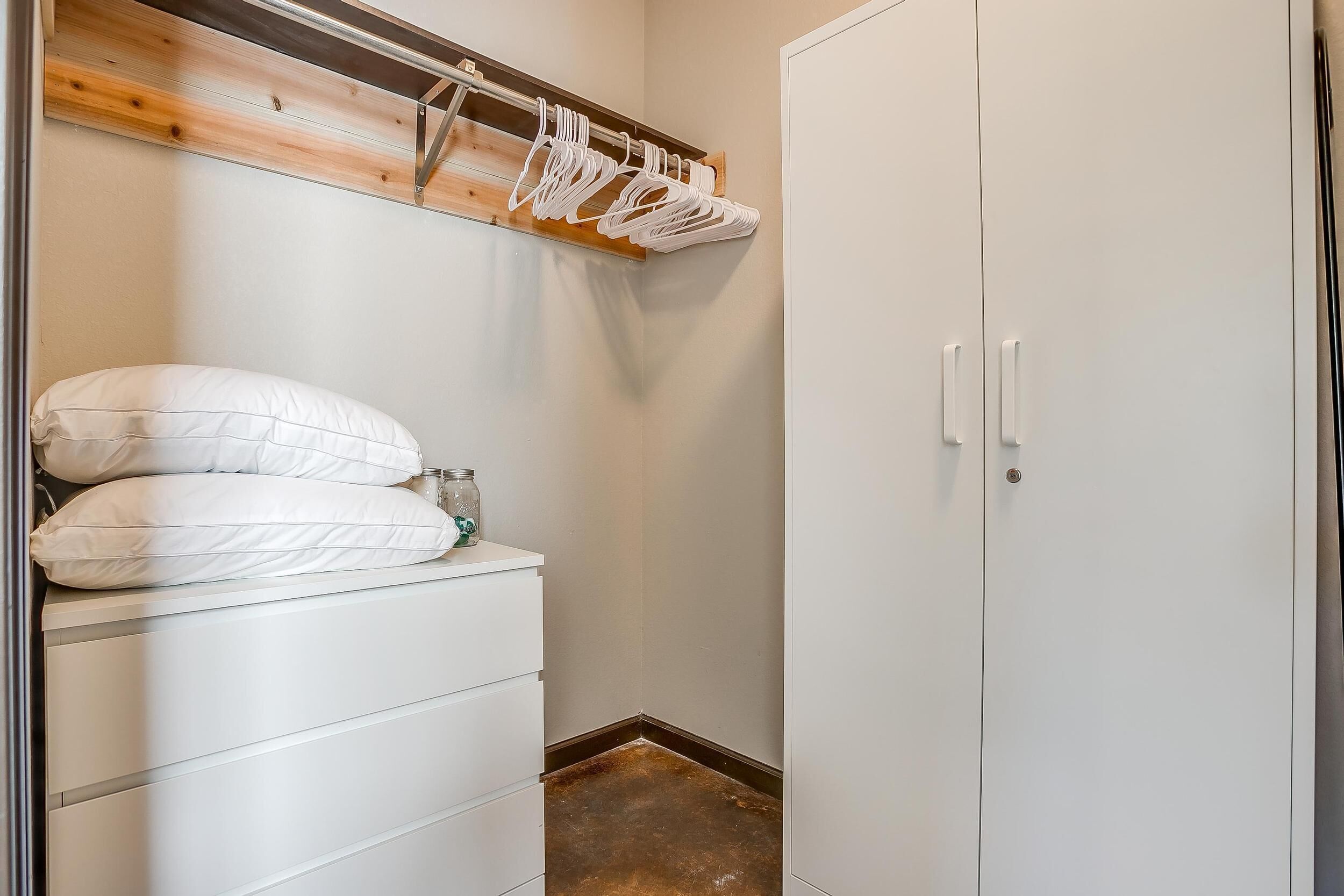 Un piccolo armadio ben organizzato con ripiano in legno sopra il letto, un letto bianco con due cuscini e un guardaroba bianco con due ante.