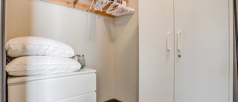 Un piccolo armadio ben organizzato con ripiano in legno sopra il letto, un letto bianco con due cuscini e un guardaroba bianco con due ante.