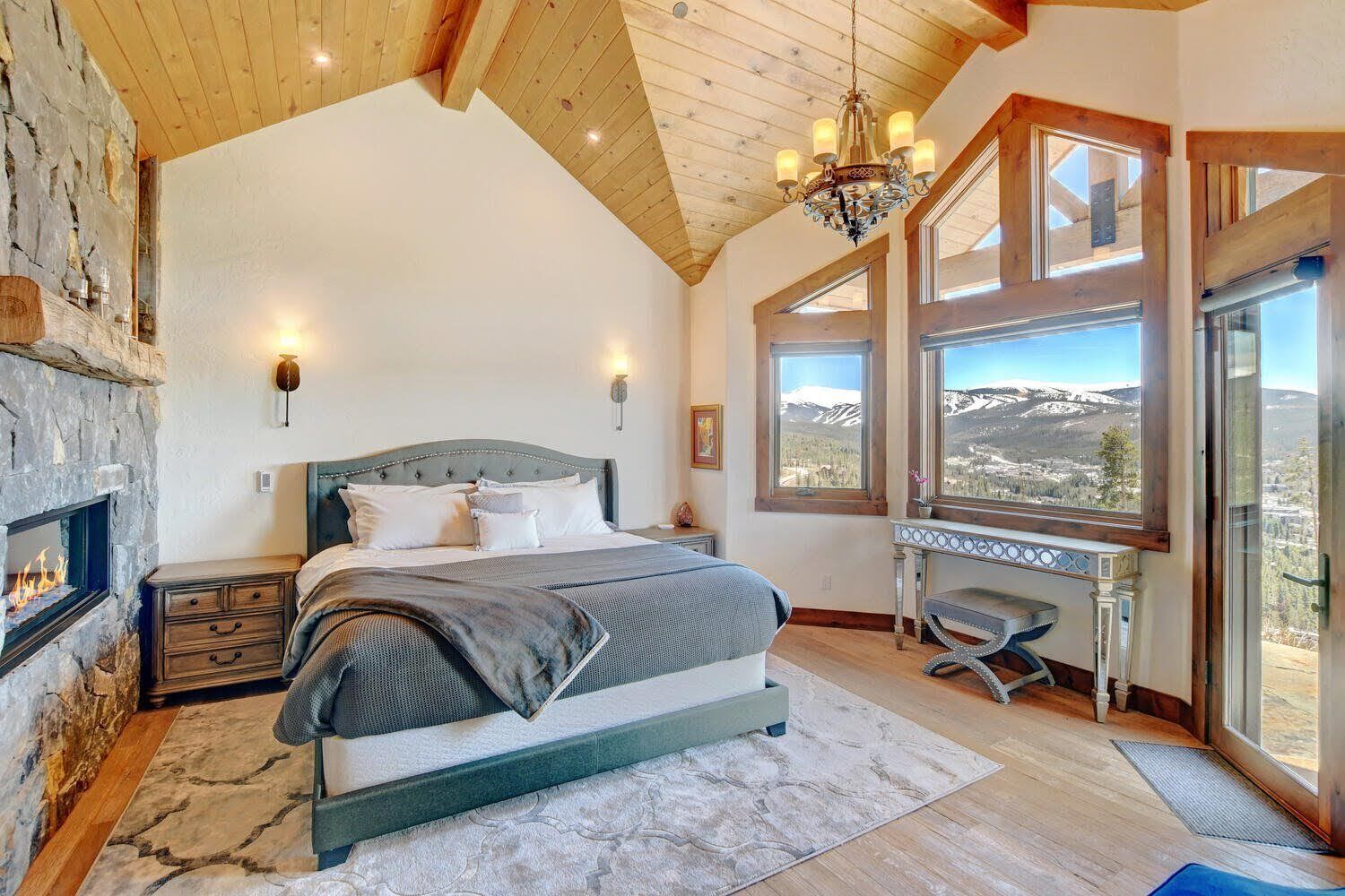 A bedroom with a large bed, a stone fireplace, a desk with a view, and a chandelier.