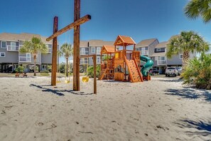 A beachside playground with a slide and swings, surrounded by palm trees and a residential building in the background.