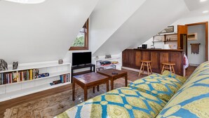 Un espace de vie de style loft avec un plancher en bois, un canapé recouvert d’un tissu à motifs, un téléviseur, une bibliothèque et un coin cuisine avec des tabourets.