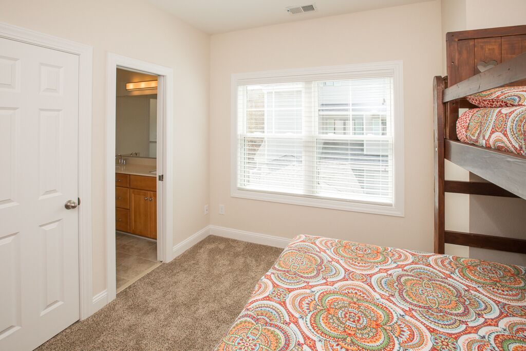 A bedroom with a bunk bed, a window with blinds, a door leading to a bathroom, and a carpeted floor.