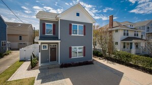 A two-story residential building with a grey exterior and red shutters, a front entrance, and a driveway leading to a gated parking area. - Beach Music - North End VB (Virginia Beach)