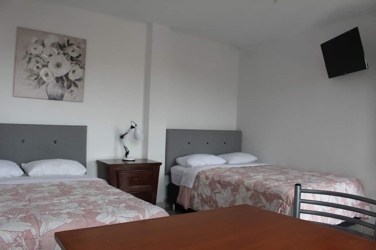 A hotel room with two beds, a wooden table, a nightstand, a lamp, a painting, and a TV mounted on the wall.