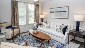 A living room with a white sofa, a wooden coffee table, a ceiling fan, and a large window with curtains. - Walk to the square and the grove! This 2BR 2BA is the perfect location.  (Oxford)