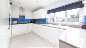 A modern kitchen with white cabinets, a blue backsplash, and a window with a view of the outdoors. - Stunning 4-Bed House in Gidea Park (Romford)
