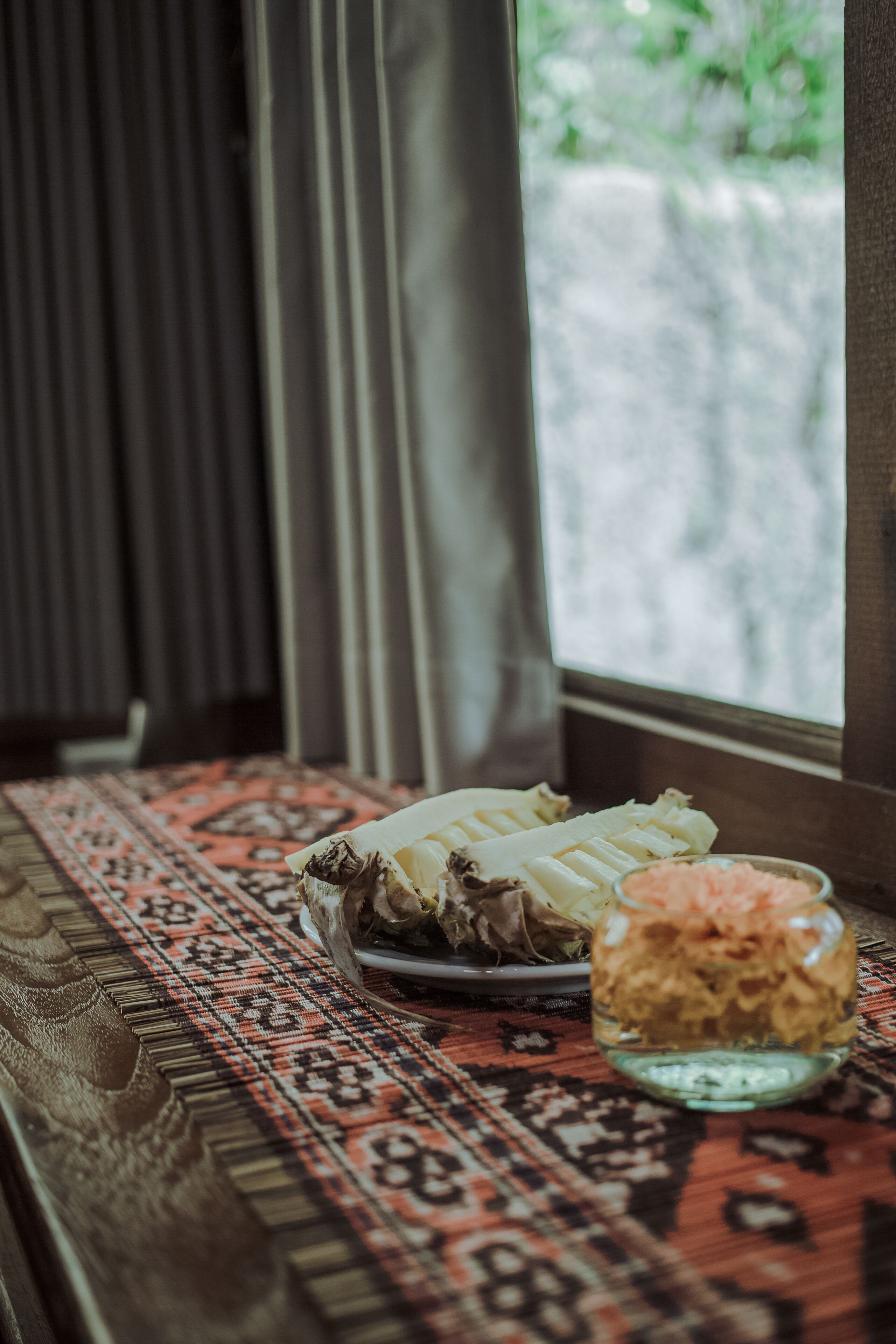 A table with a plate of food and a glass of sauce, set on a patterned rug near a window with curtains.
