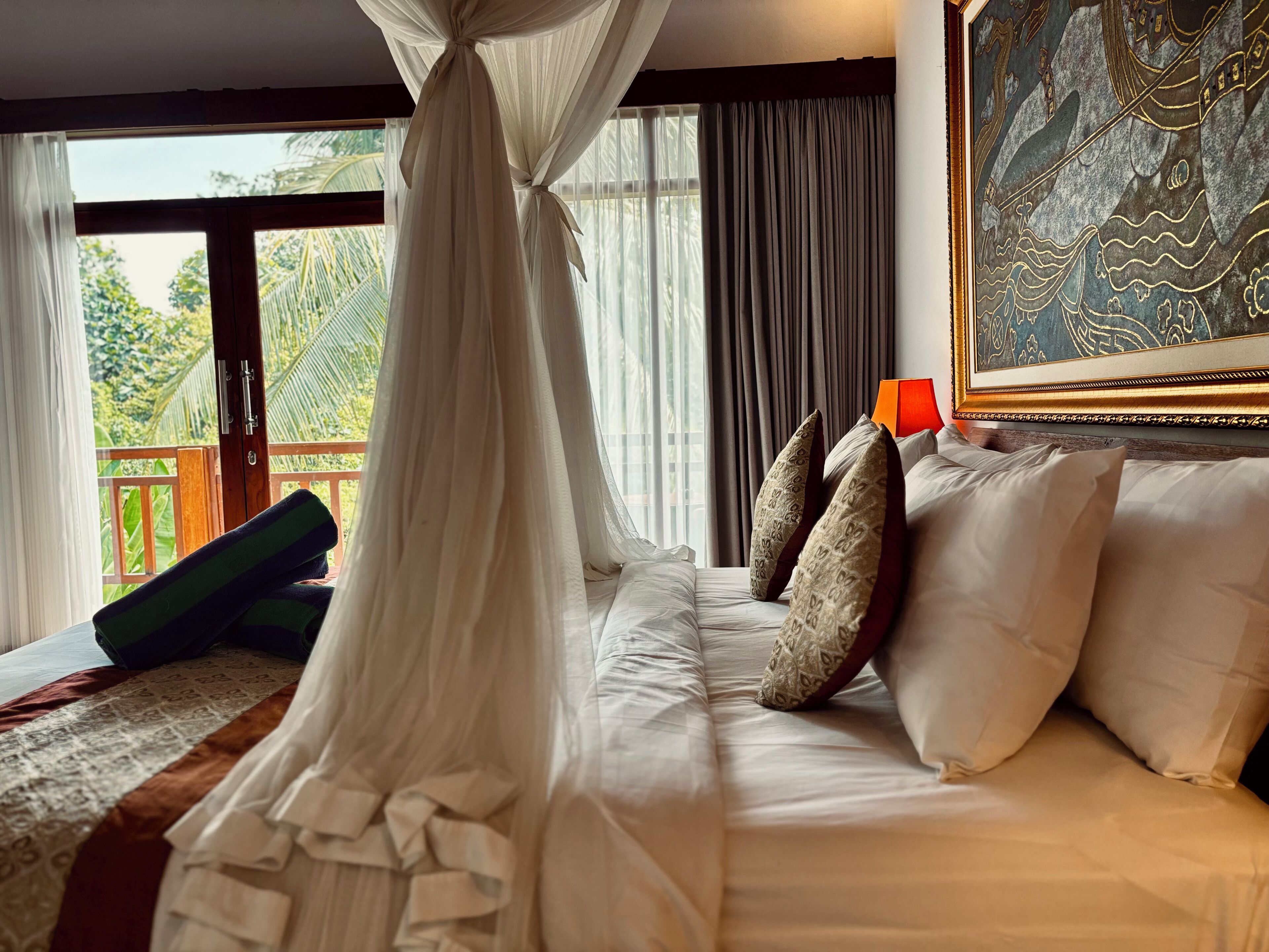 A bedroom with a bed, pillows, a canopy, a painting, and a view of greenery.
