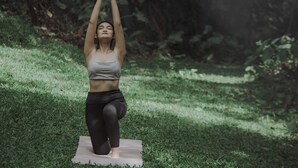 A person performing a yoga pose on a mat in a grassy outdoor area with trees in the background.