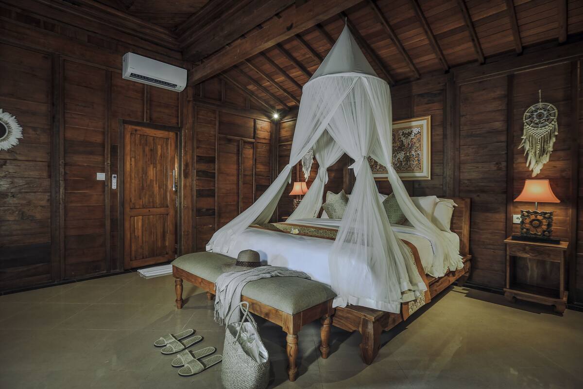 Une pièce en bois avec un lit sous un dais, un banc, une table de chevet avec une lampe et un appareil de climatisation.