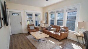 A living room with a brown leather sofa, a wooden coffee table, and a TV mounted on the wall. - Brand New Craftsman 2BR 2BA Home in Denver Heights (San Antonio)