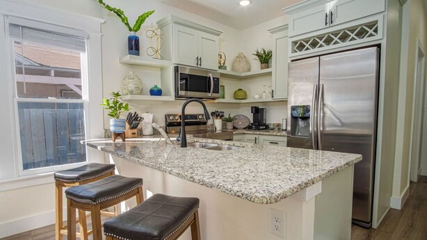 A modern kitchen with white cabinetry, a granite countertop, stainless steel appliances, and wooden stools. - Brand New Craftsman 2BR 2BA Home in Denver Heights (San Antonio)