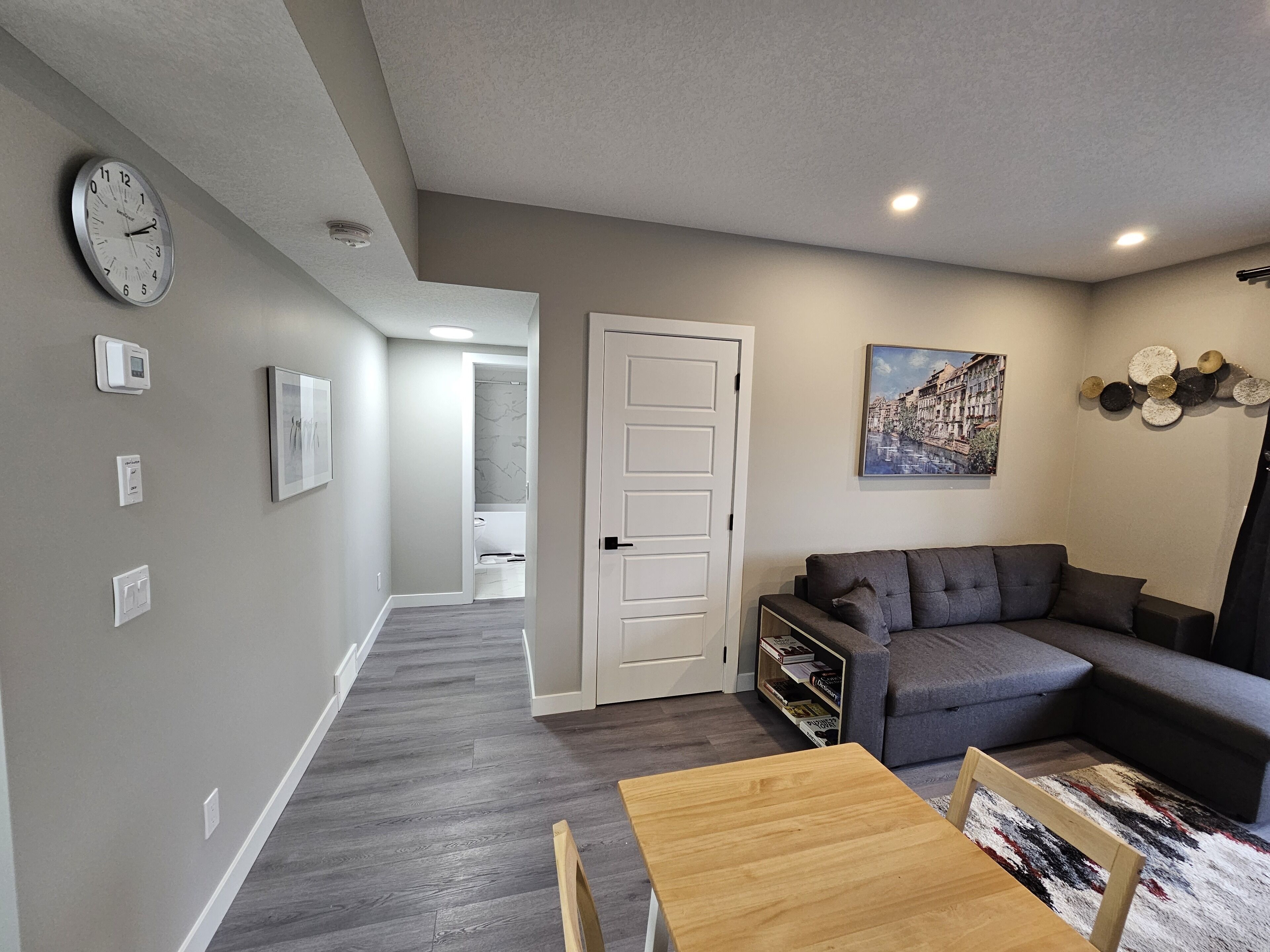 A modern living room with a grey sofa, a wooden coffee table, and a dining table. There is a white door leading to another room, and a clock on the wall.