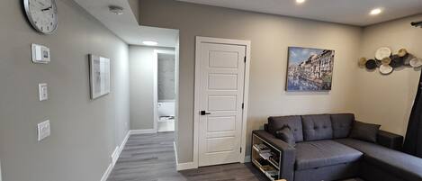 A modern living room with a grey sofa, a wooden coffee table, and a dining table. There is a white door leading to another room, and a clock on the wall.