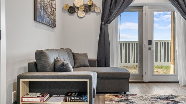 A modern living room with a grey sectional sofa, a wooden side table with books, a decorative wall piece, and a view through a glass door to an outdoor deck. - Sunny Walk-out Unit | Free HiSpeed WiFi & Parking | Near Grocery (Calgary)