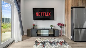 A modern living room with a large flat-screen TV mounted on the wall, a dark wooden cabinet, a stainless steel refrigerator, and a large window with curtains. - Sunny Walk-out Unit | Free HiSpeed WiFi & Parking | Near Grocery (Calgary)