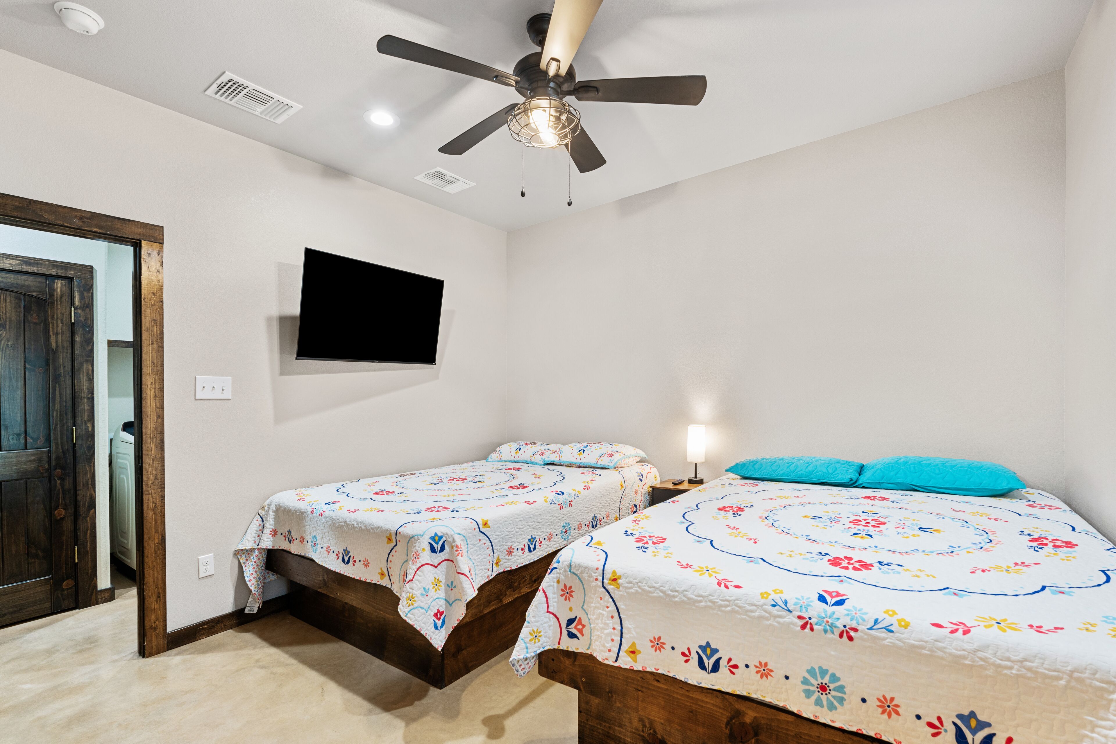 A room with two wooden beds, a ceiling fan, a mounted TV, and a door.