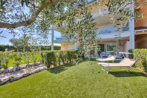Un espace extérieur moderne avec une pelouse verdoyante, une pergola ornée d’oliviers et un fauteuil de relaxation.