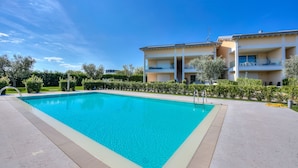 A rectangular swimming pool with a tiled deck, surrounded by a well-maintained hedge and a multi-story building with balconies.
