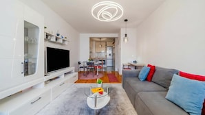 Un salon moderne avec un téléviseur à écran plat, une table basse en verre, un canapé gris agrémenté de coussins rouges et bleus, et une cuisine en arrière-plan.