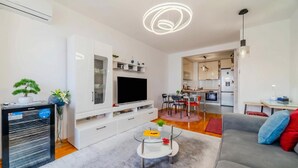 Un salon moderne avec un téléviseur à écran plat, une table basse en verre, un canapé gris et une cuisine en arrière-plan.
