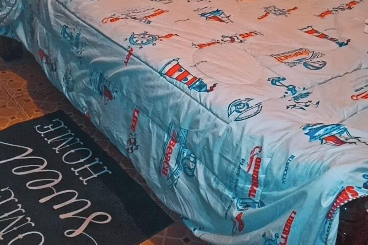 A single bed with a patterned bedspread, a pillow, and a black mat with text on the floor.