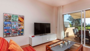 A modern living room with a flat-screen TV on a white cabinet, a glass coffee table, and a sofa with striped cushions.