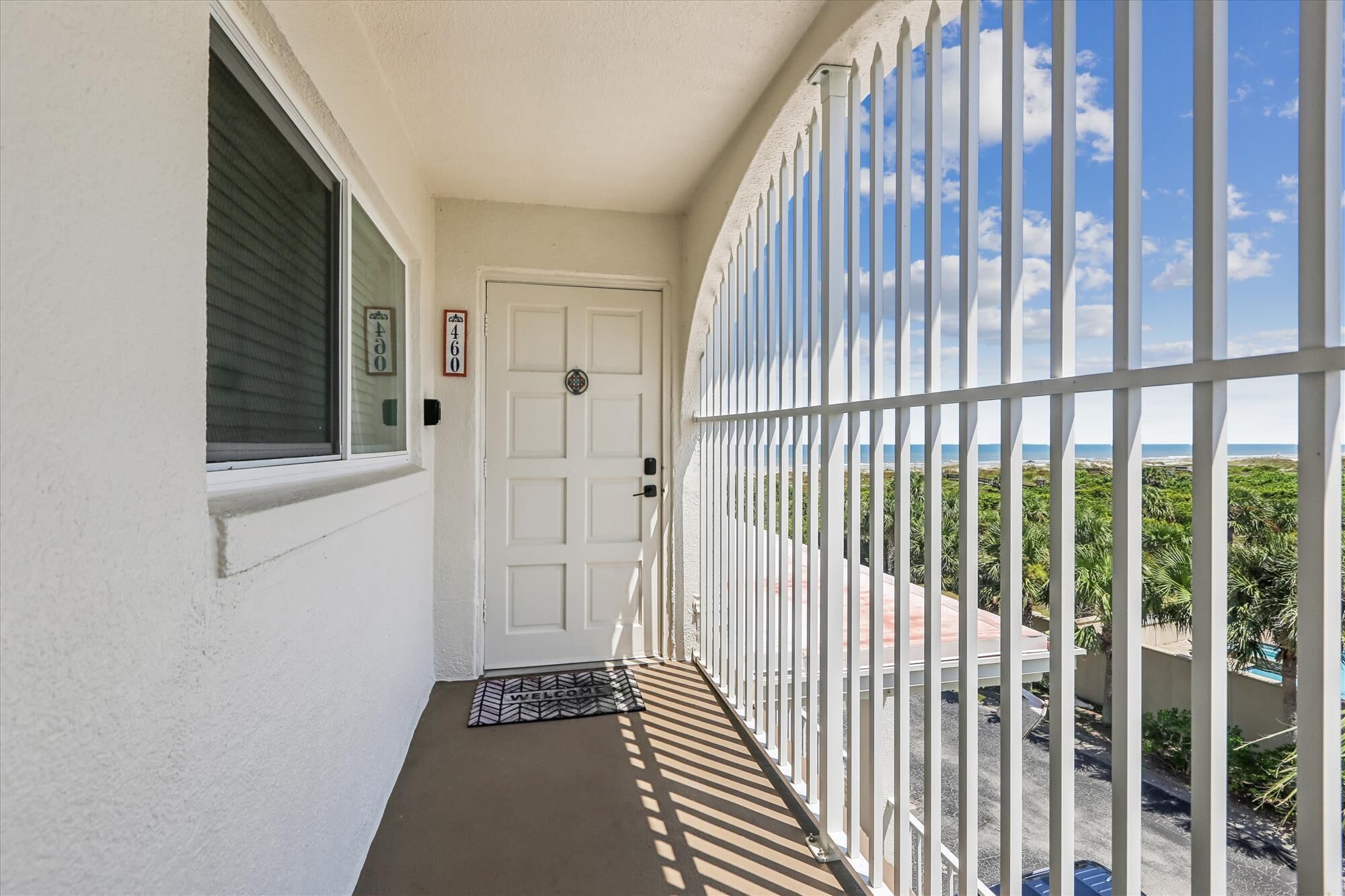 A narrow balcony with a white door, a window with blinds, and a view of a beach with greenery.
