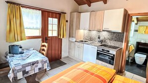 Ein gemütlicher Raum mit Bett, Tisch mit Tischdecke, Stuhl, einer kleinen Küchenzeile und einem Fenster mit Vorhängen.