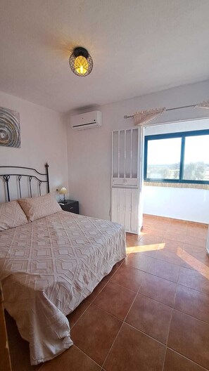 Ein Schlafzimmer mit Bett, Klimaanlage, einem Fenster mit Vorhängen und Blick ins Freie.