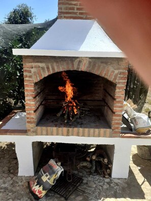 Ein gemauerter Außenofen mit weißer Fassade und sichtbarem Feuer darin.