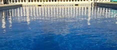 A swimming pool with a white balustrade, clear blue water, and a partly cloudy sky.