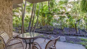 A covered outdoor seating area with a round glass table and metal chairs, surrounded by lush tropical plants and palm trees.