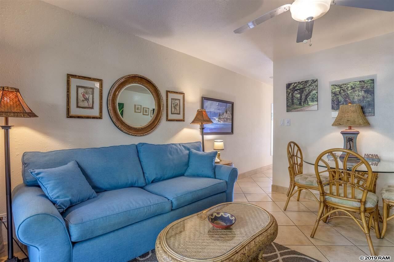 A living room with a blue sofa, a round mirror, framed pictures, a dining table with chairs, and a ceiling fan.