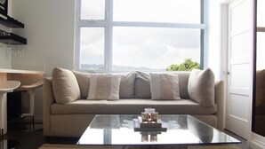 A modern living room with a beige sofa, a glass coffee table, and a view through a large window.